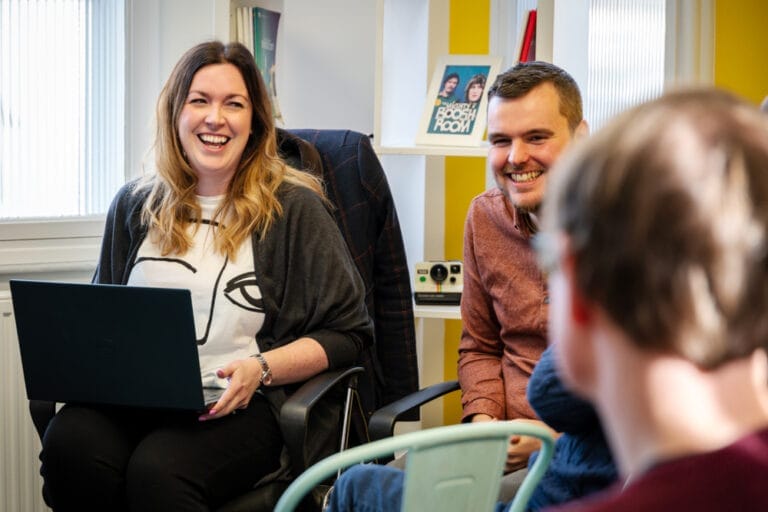 Two colleagues laugh during an informal office meeting, one using a laptop while a third person sits in the foreground, with shelves and a retro instant camera in a bright workspace behind them.