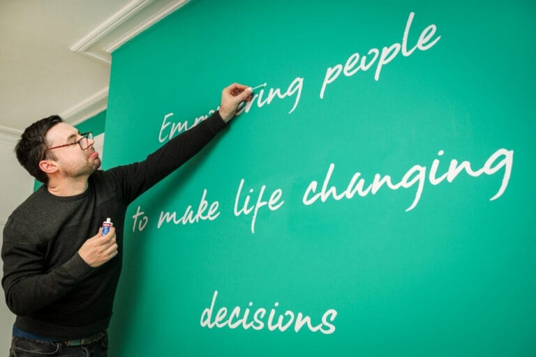 Man with glasses writes a white cursive wall slogan on a teal wall reading “Empowering people to make life changing decisions.”