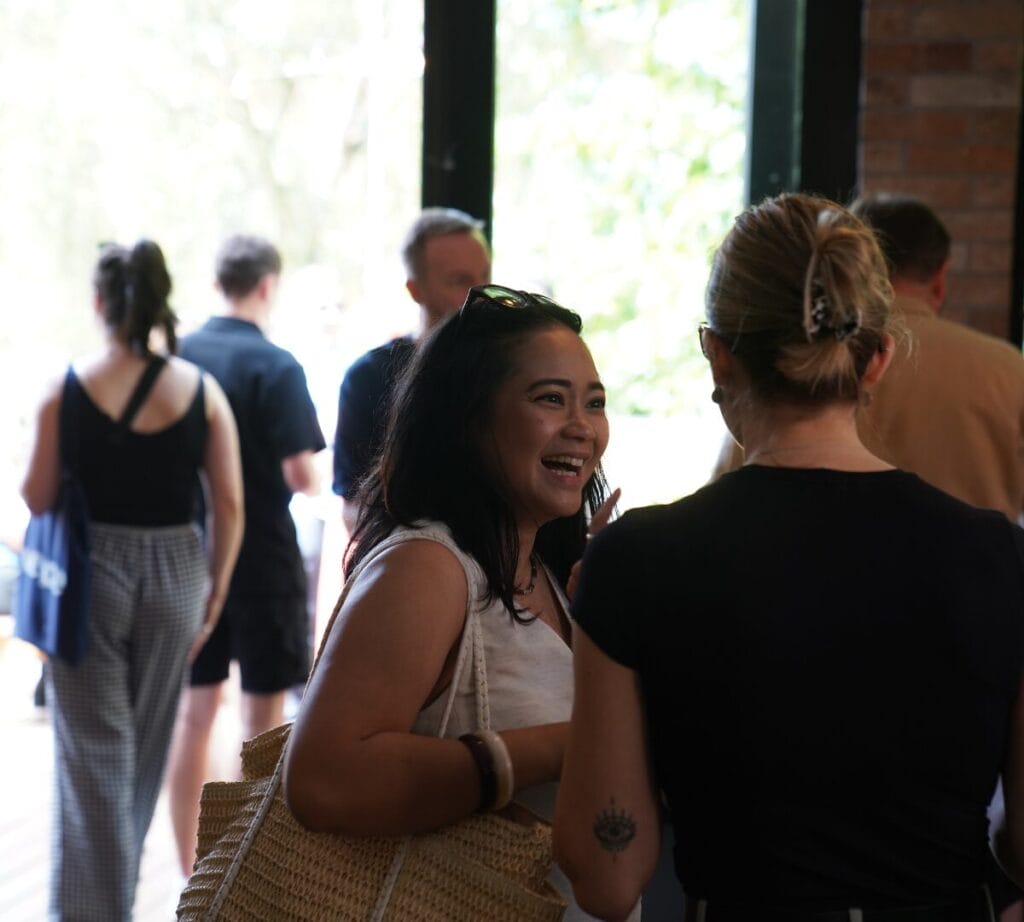 A woman is smiling at her friend or colleague, as they gather in the briefing room of the Vepple Team-Building Murder Mystery
