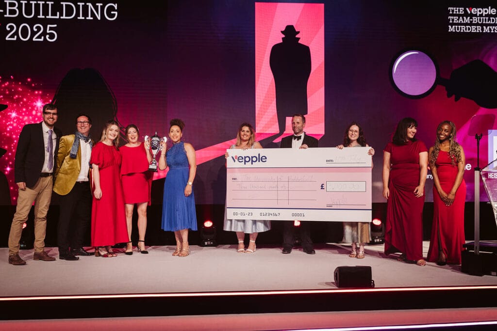 The Vepple team are on stage, with the University of Huddersfield winners clutching their trophy and Vepple staff holding up a giant cheque for £3000. The Vepple Team-Building Murder Mystery is in the background on the screen.