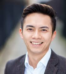 Smiling person with neatly styled dark hair in a grey blazer over a light shirt, captured in a blurred outdoor setting.