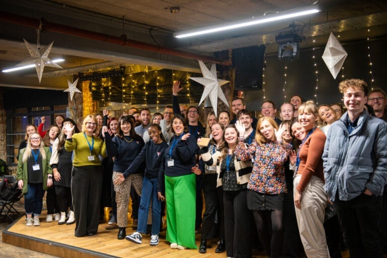 Large group of adults smiling and waving on a small stage at an indoor event, many wearing lanyards, with hanging star lanterns and fairy lights creating a festive backdrop.