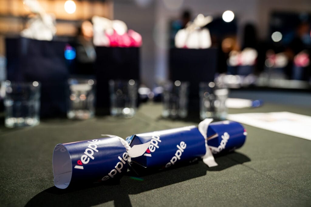A blue Christmas cracker tied with white ribbon rests on a dark table, with glass tumblers and gift bags blurred in the background at an event.