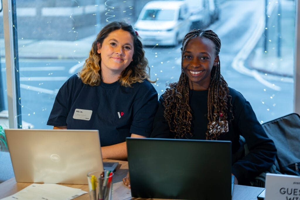 Two smiling people sit behind laptops at a window-side table, wearing dark T-shirts and name badges, with fairy lights and a street outside and a small “Guest” sign on the desk.