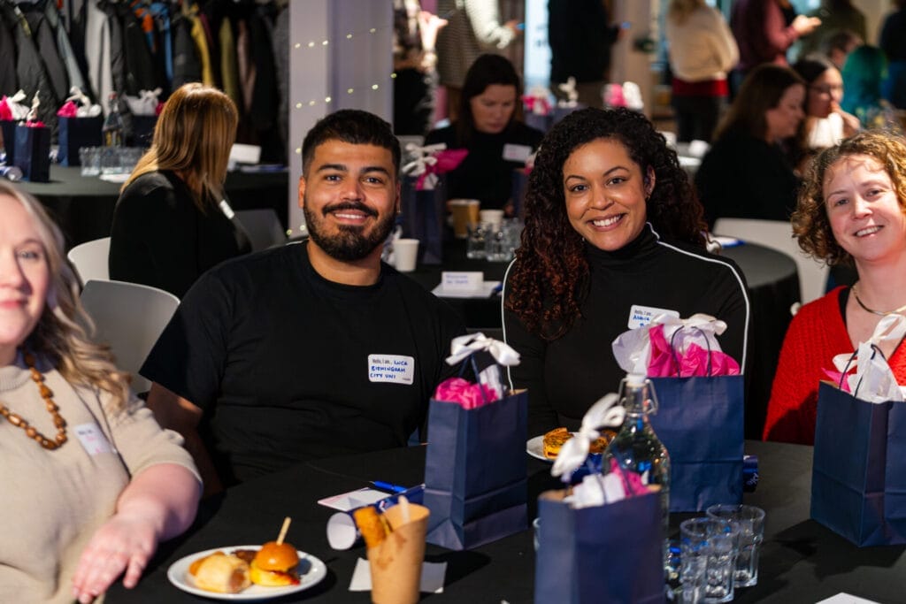 Smiling attendees sit at a round table during a social event, wearing name tags and surrounded by blue gift bags with pink tissue paper, drinks, and small plates of snacks.