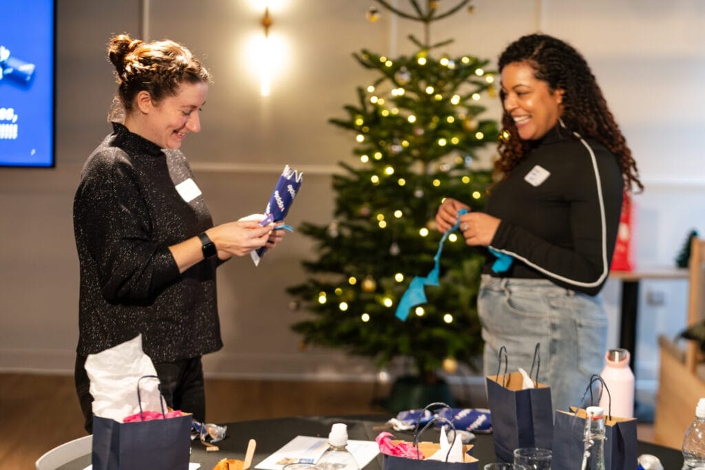 Two colleagues smile while opening festive crackers at a table with gift bags and snacks, with a lit Christmas tree in the background.