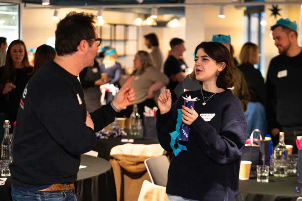 Two people with name badges chat animatedly at a casual indoor event, one gesturing as the other holds a blue party cracker, with other attendees and tables with drinks in the background.