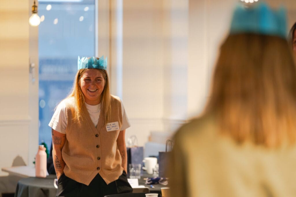 Smiling person in a blue paper crown and beige knitted vest stands by a table at a casual indoor gathering, with another crown-wearing guest blurred in the foreground and gift bags and glasses on the table.