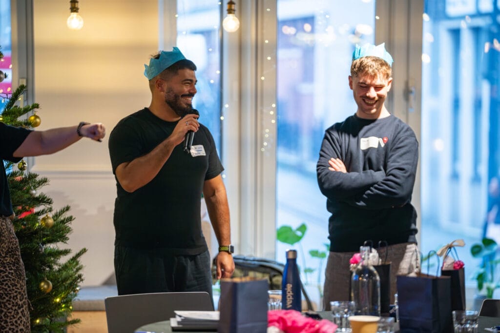 Two people wearing blue paper crowns share a laugh as one speaks into a microphone at a festive indoor gathering, with a decorated Christmas tree and tables with gift bags and drinks in the foreground.