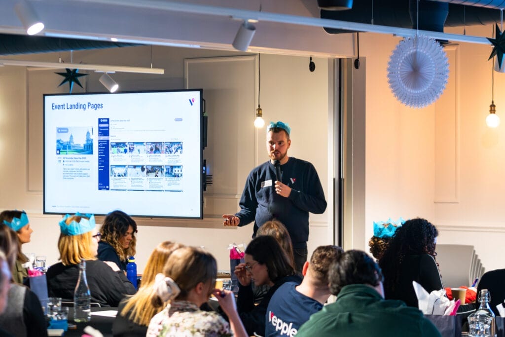 A speaker with a microphone presents a slide titled “Event Landing Pages” on a large screen to a seated audience in a modern room with festive decorations, including paper crowns and hanging lights.