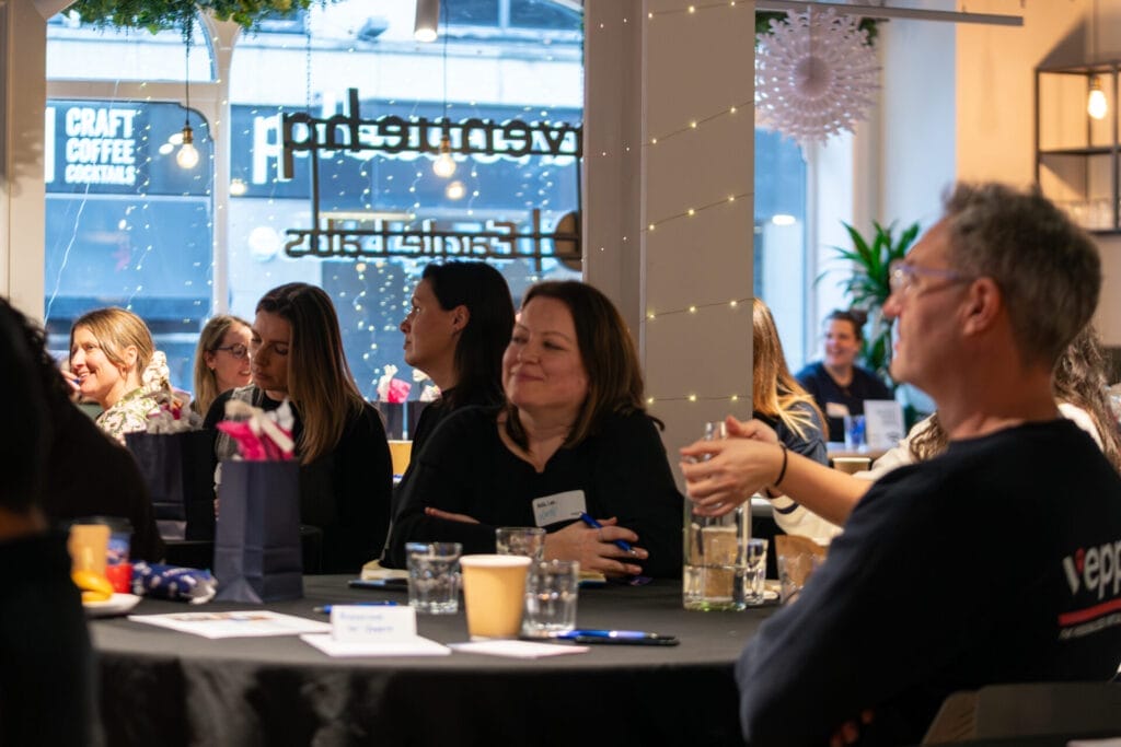 Participants sit around tables at an informal indoor event, with cups, notebooks and gift bags, while warm fairy lights and a “Craft Coffee Cocktails” window sign glow behind.