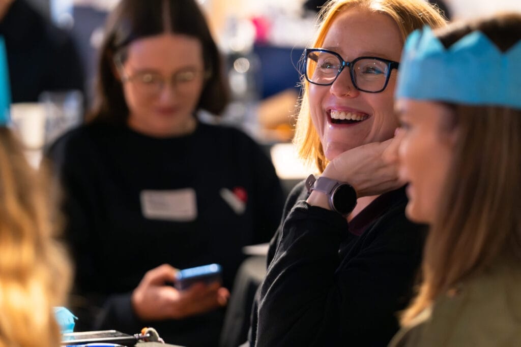 Smiling person with glasses and a smartwatch laughs with others at a gathering, with blurred attendees behind, one using a phone and another wearing a blue paper crown.