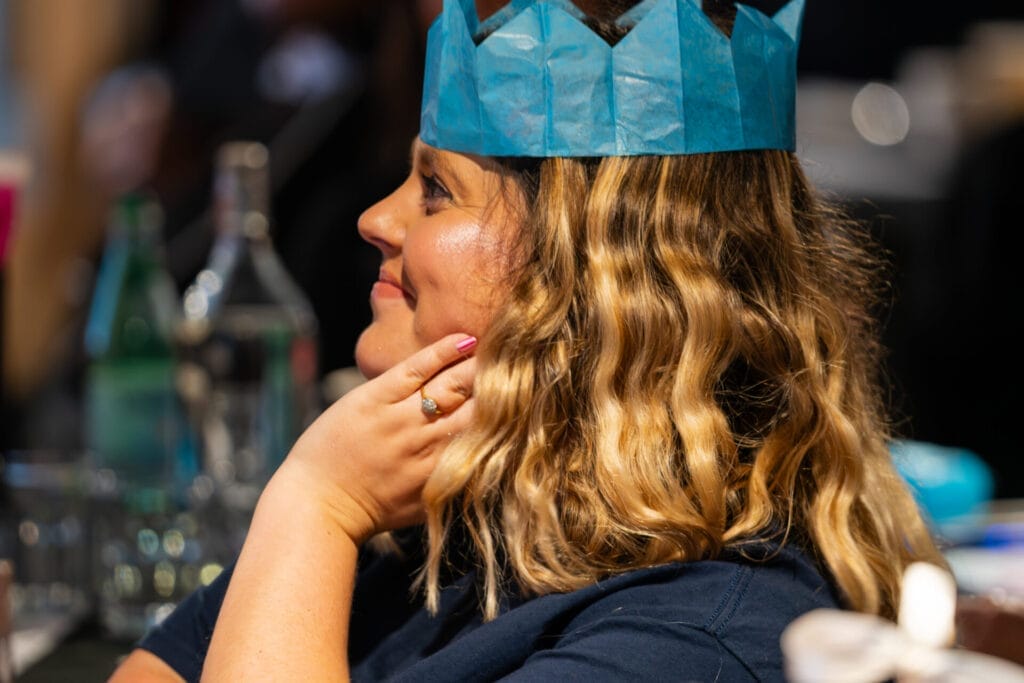Person in profile wearing a blue paper crown smiles with wavy blonde hair, resting a hand with pink nails and a ring on their cheek, with blurred bottles and glasses in the background.