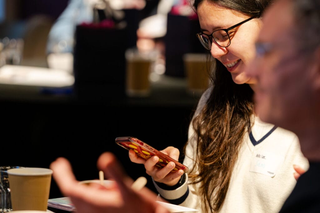 A person with long dark hair and glasses smiles at a smartphone at a table during an indoor event, with coffee cups and a blurred attendee in the foreground.