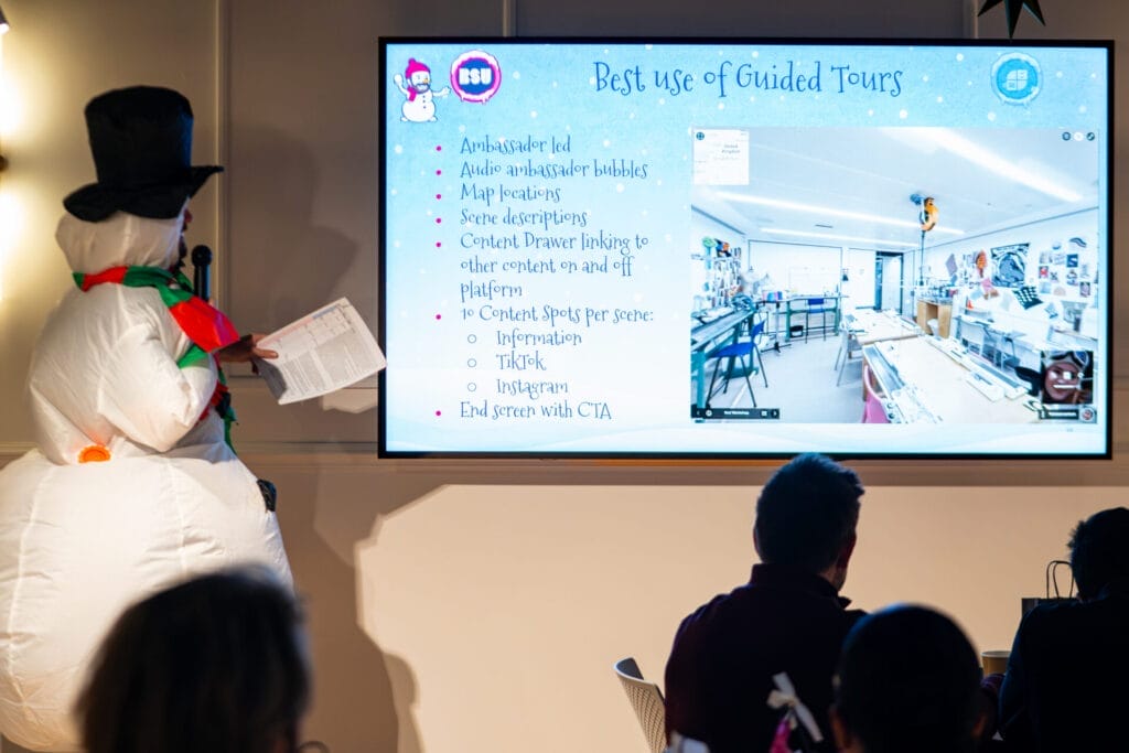 A presenter in a snowman costume stands beside a large screen showing a slide titled Best use of Guided Tours with bullet points and a classroom virtual tour, as an audience watches.
