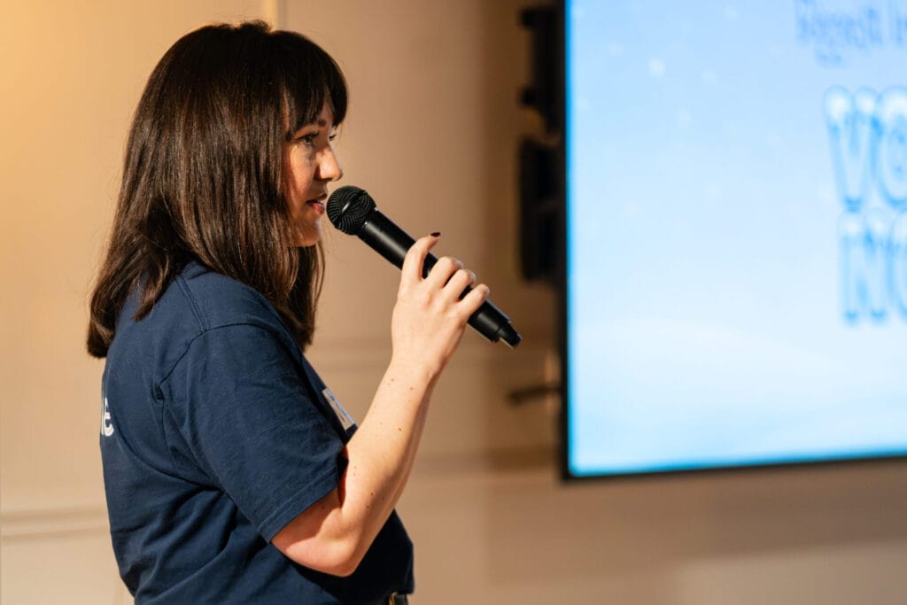 Side-profile of a speaker with shoulder-length dark hair in a navy T-shirt holding a microphone beside a large presentation screen.