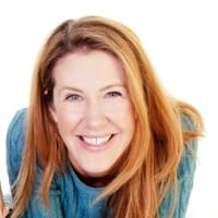 A smiling person with long strawberry-blonde hair wears a teal jumper on a white background.