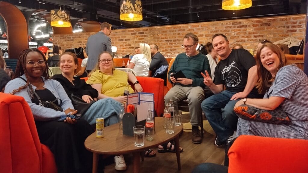 Six friends relax on orange sofas in a cosy bar, smiling at the camera with drinks and menus on a small table between them.