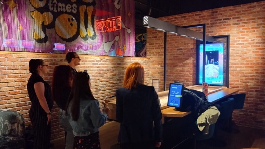 People watching a shuffleboard-style game on a digital screen in a neon-lit bar with brick walls and arcade-style decor.
